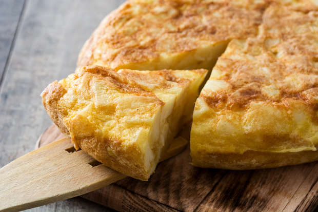 A slice of golden-brown tortilla is being lifted from a wooden board with a spatula, surrounded by a rustic, wooden surface.